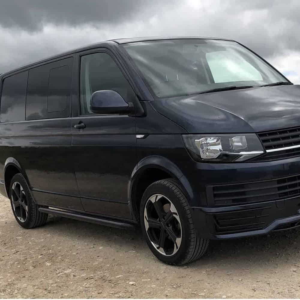 Black VW T5 T6 van with side bars parked outdoors under cloudy sky.