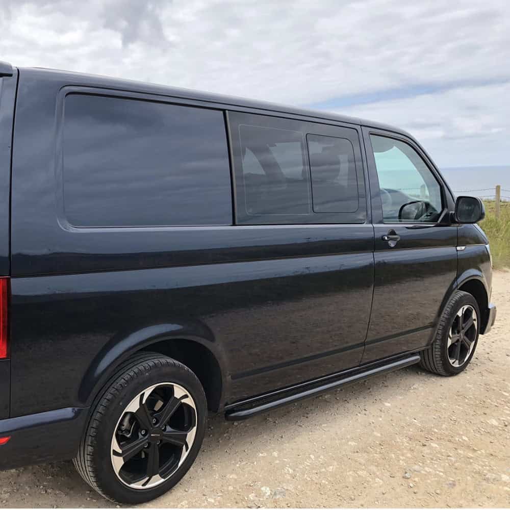 Black VW T5 T6 van with side bars parked on a gravel surface under cloudy sky.